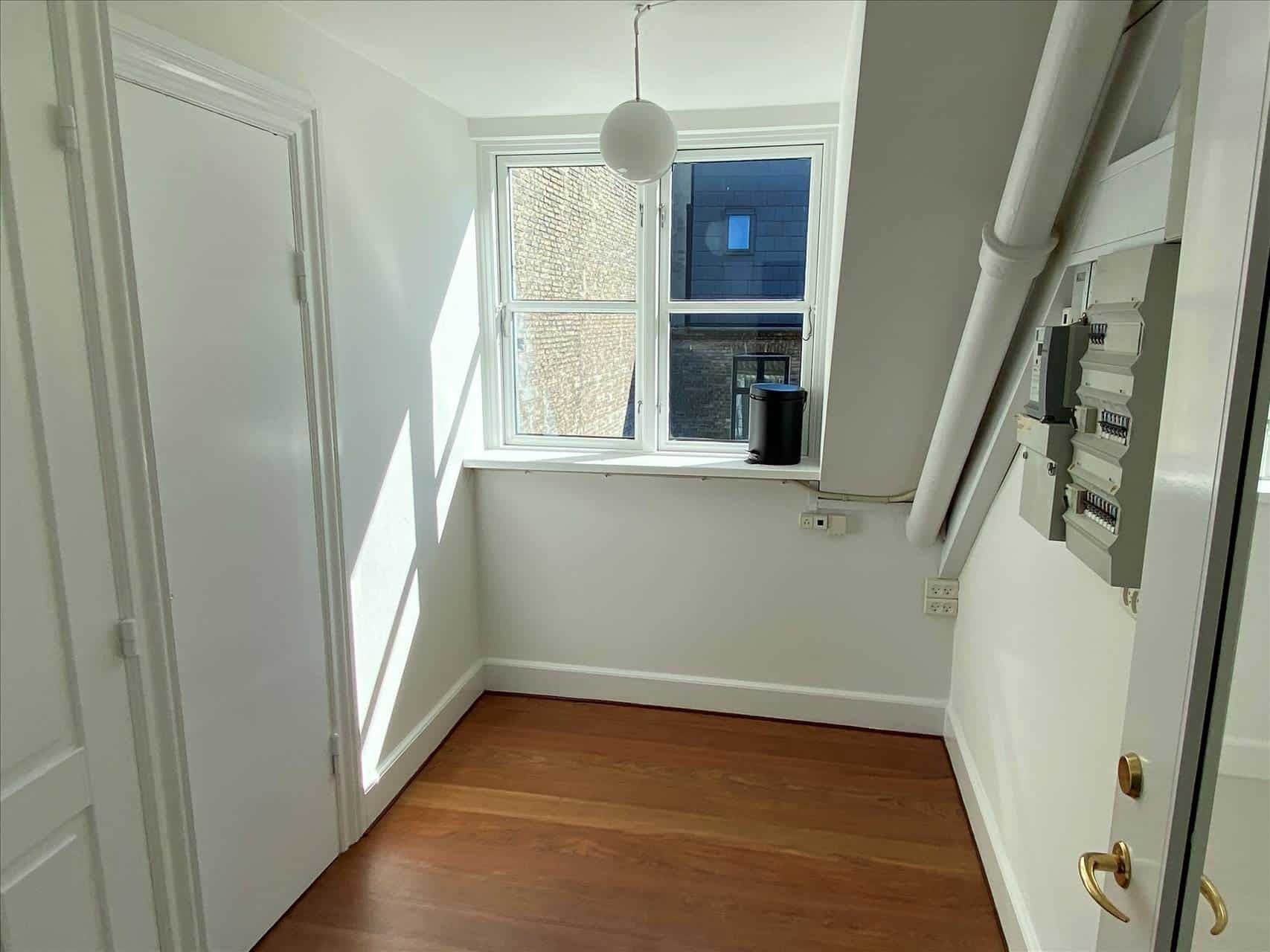 Lyst og rent kontor med trægulv og hvide vægge samt et stort vindue med udsigt over byens tage