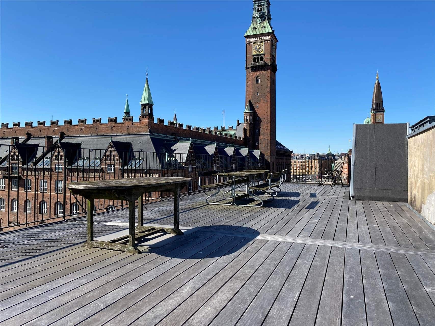 Terrasse med tre træborde og to bænke. Et klokketårn og en bygning med mange vinduer i baggrunden.