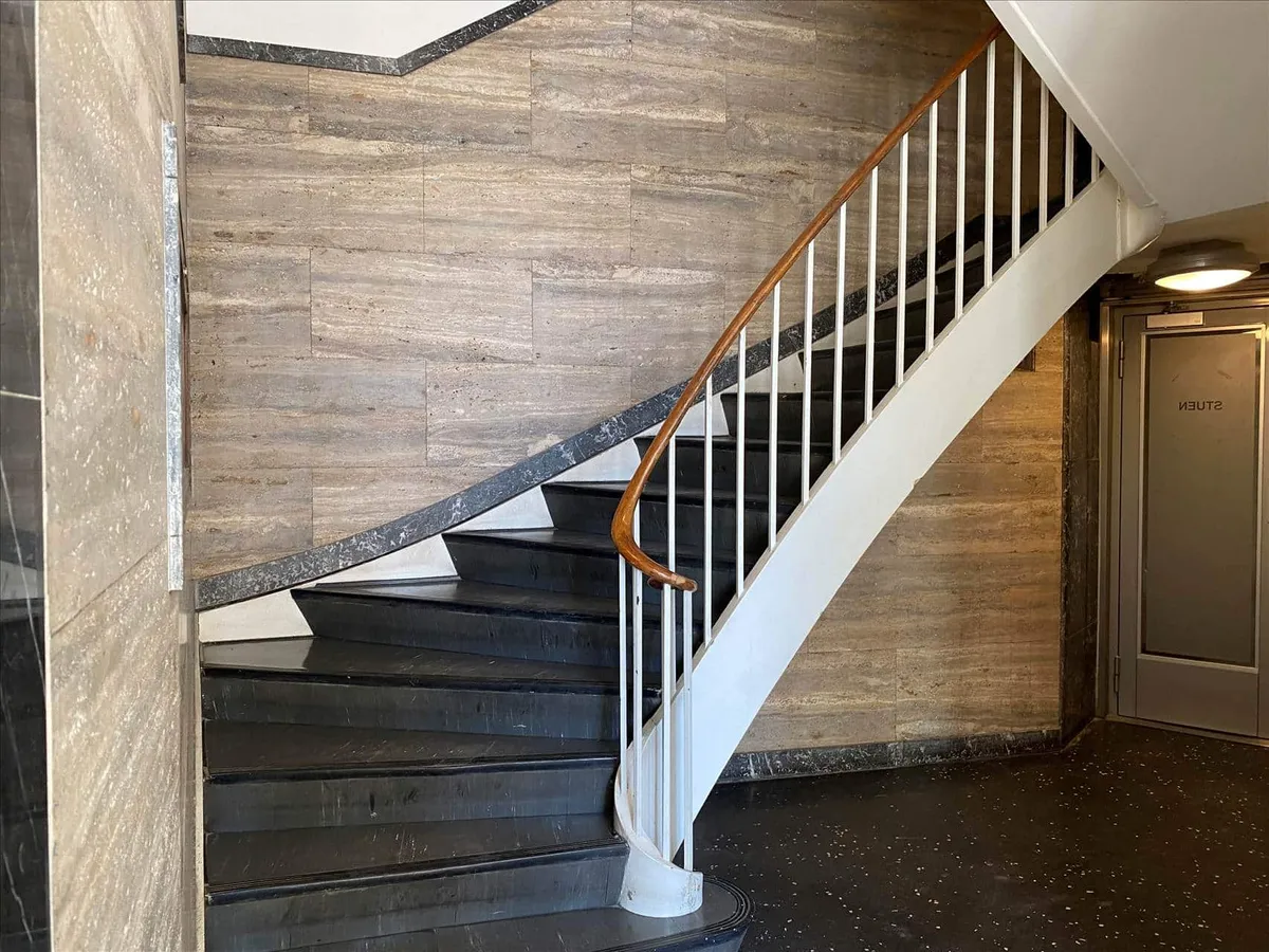 Moderne trappe med træhåndtag og hvide trappeskinner op til 2. sal, marmorgulv og lysegrå vægge med tekstur.