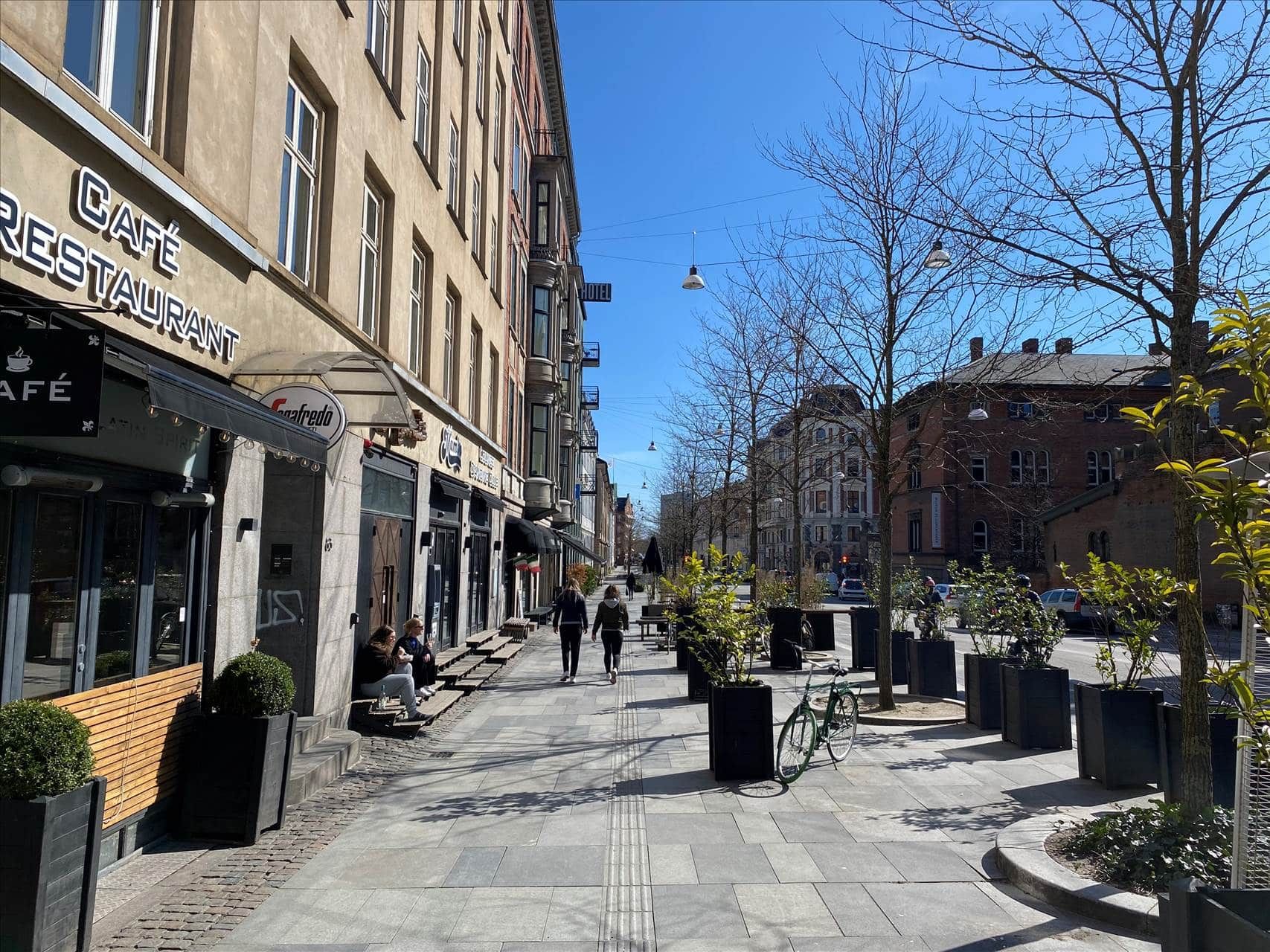 Mennesker går på en gågade med restauranter og grønne planter og træer langs vejen