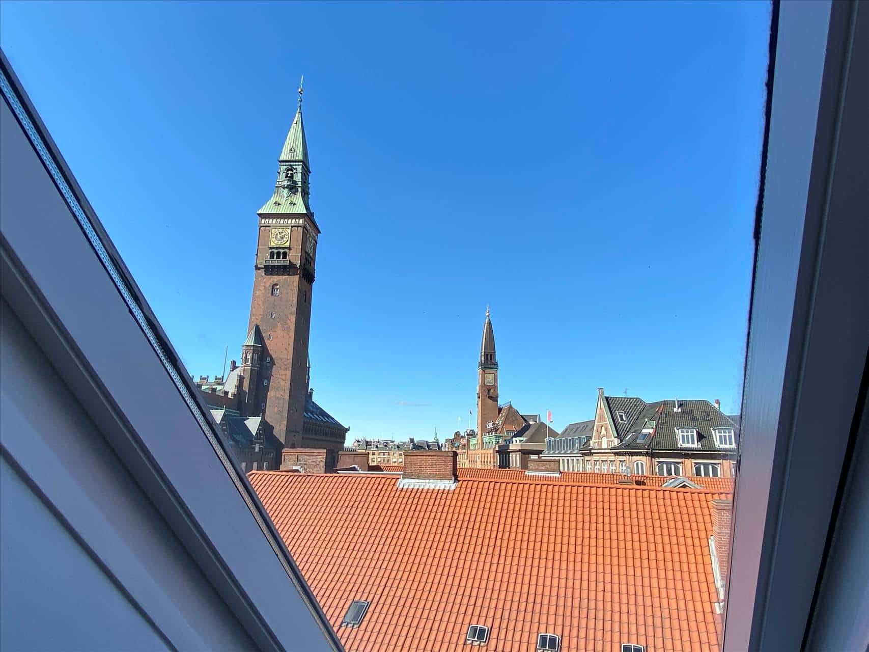 Udsigt fra et vindue over byen med røde tag og tårn med ur. Bygninger i baggrunden, blå himmel.