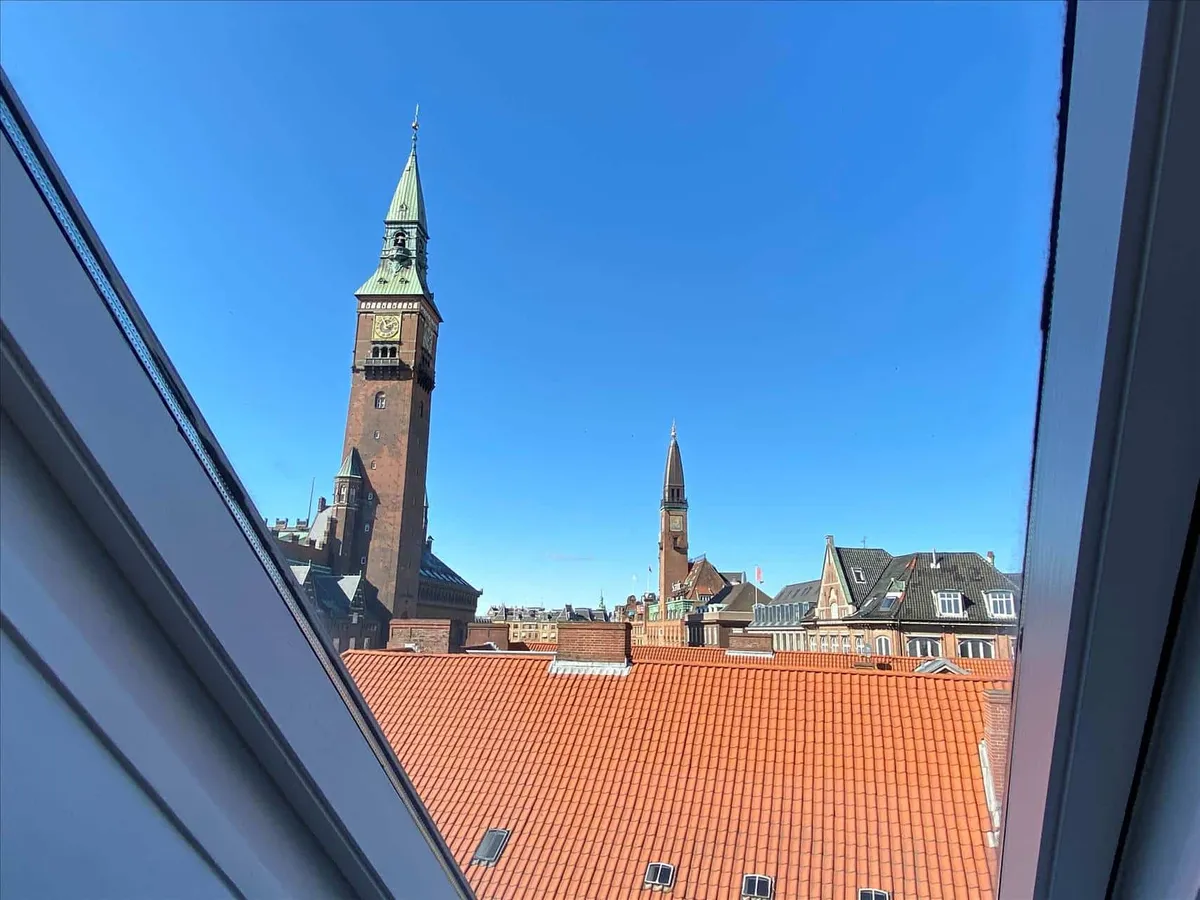 Udsigt fra et vindue over byen med røde tag og tårn med ur. Bygninger i baggrunden, blå himmel.