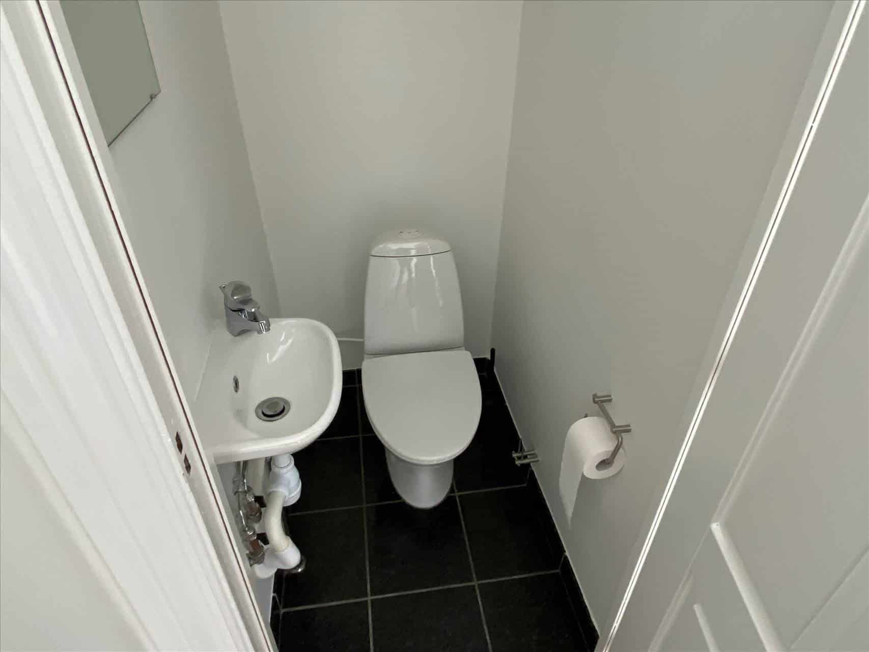 Et lille hvidt badeværelse med toilet, håndvask og toiletpapir. Toiletstol og håndvask er placeret på en sort fliseret gulv.