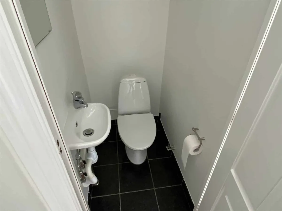 Et lille hvidt badeværelse med toilet, håndvask og toiletpapir. Toiletstol og håndvask er placeret på en sort fliseret gulv.