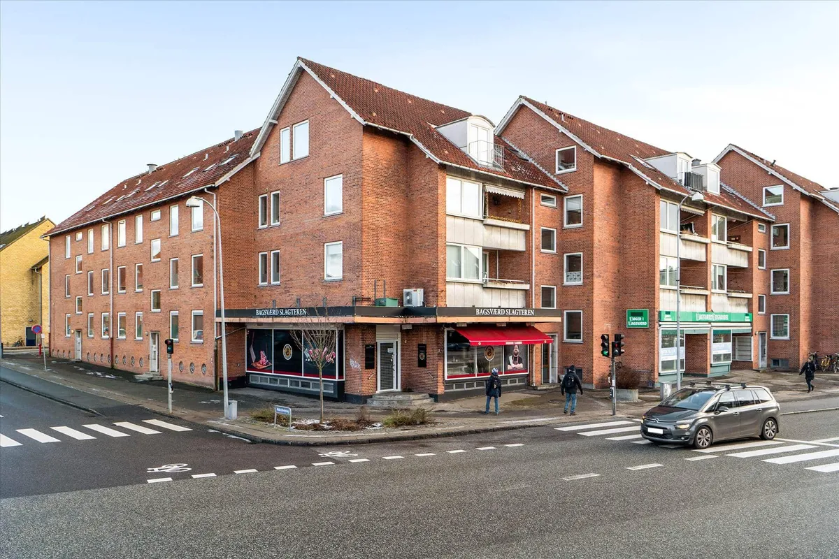 Lejlighedsbygning med røde mursten og vinduer, to butikker, fortov med gågade og mennesker og biler