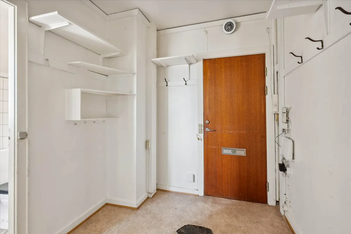 Lejlighedsgang med hvide vægge og brun dør, indgangsdør med sølvfarvet håndtag, flere hylder og et toilet på venstre side