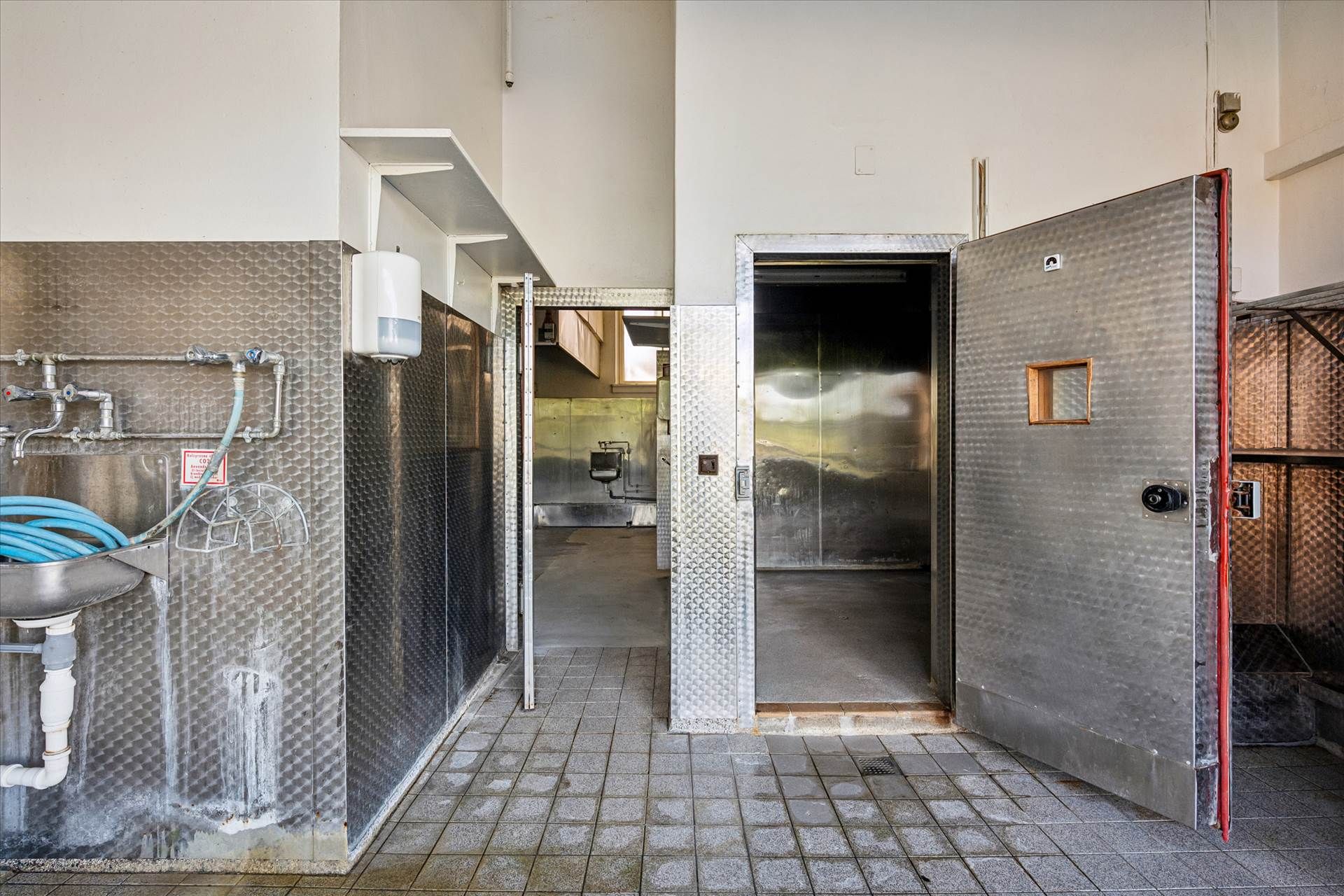 En lukket lagerdør med et lille vindue og en åben dør i en stor hall med en vaskebækken og et toilet.
