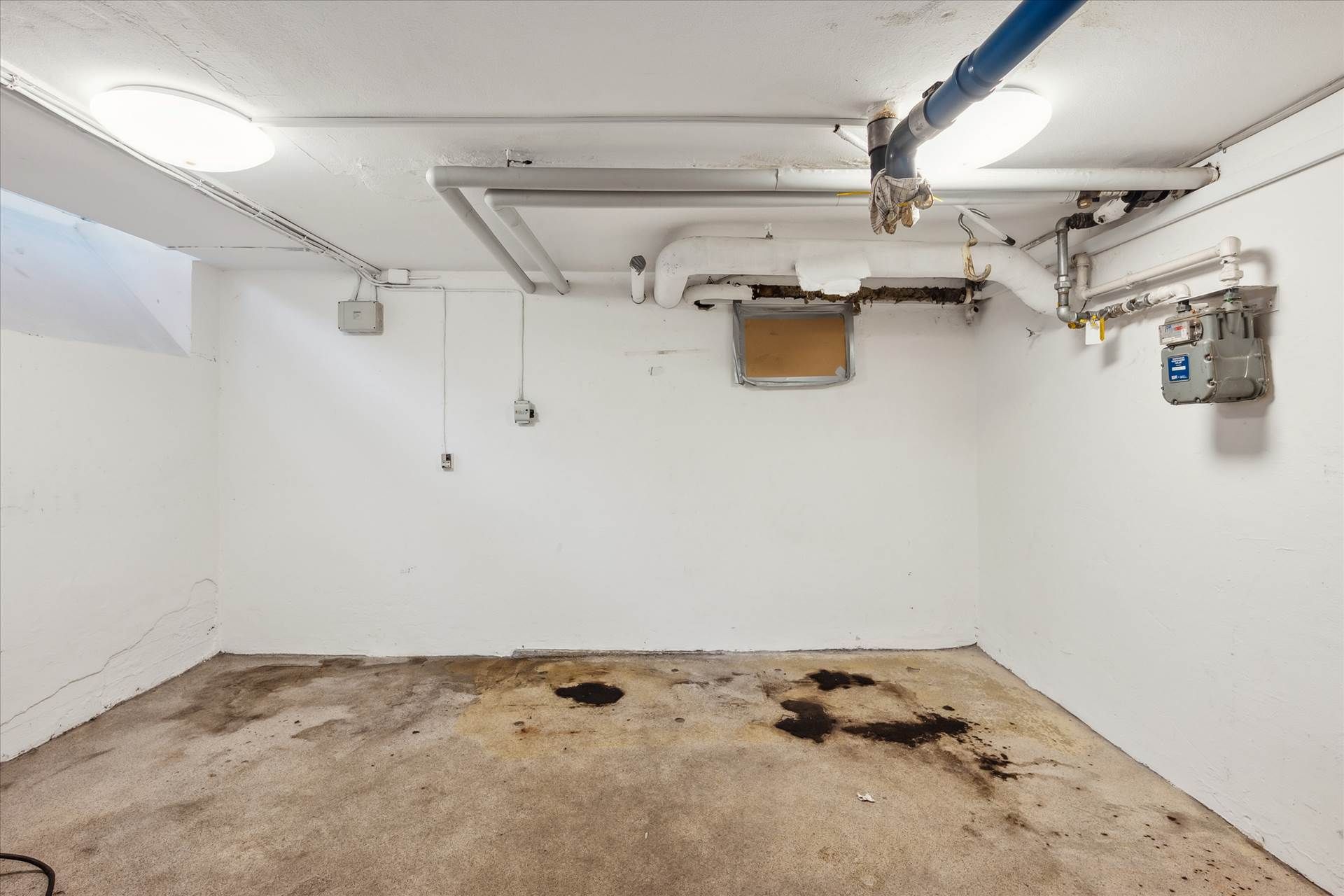 En tom garage med lyse gulvbelægning og hvide vægge. Der er to lysekroner i loftet og en meterboks samt et ventilpanel på