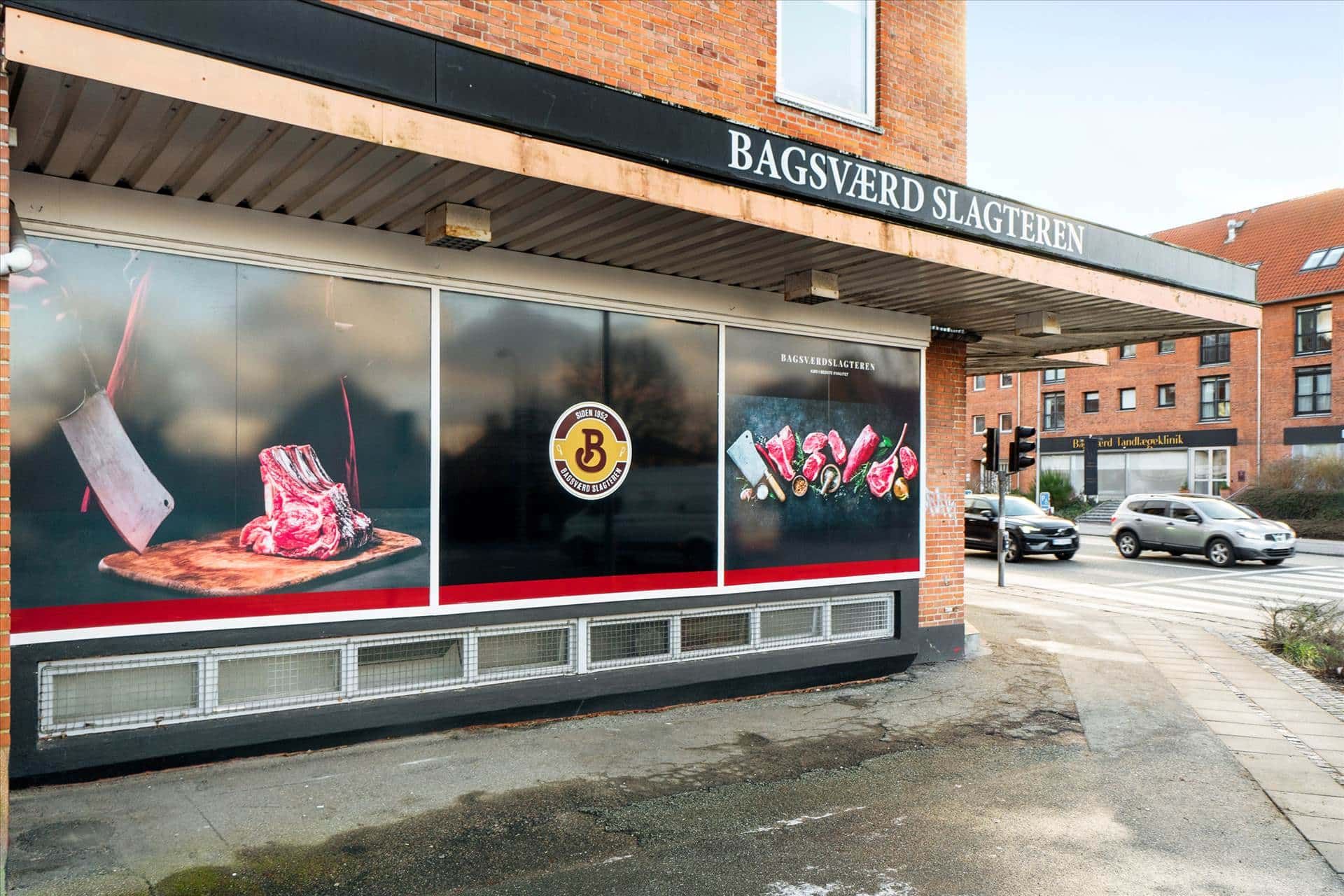 Butiksfront med udsigt til Bagsværd Slagteren. Store glasruder med reklamebilleder af rått kød. Kød og kniv på den ene side