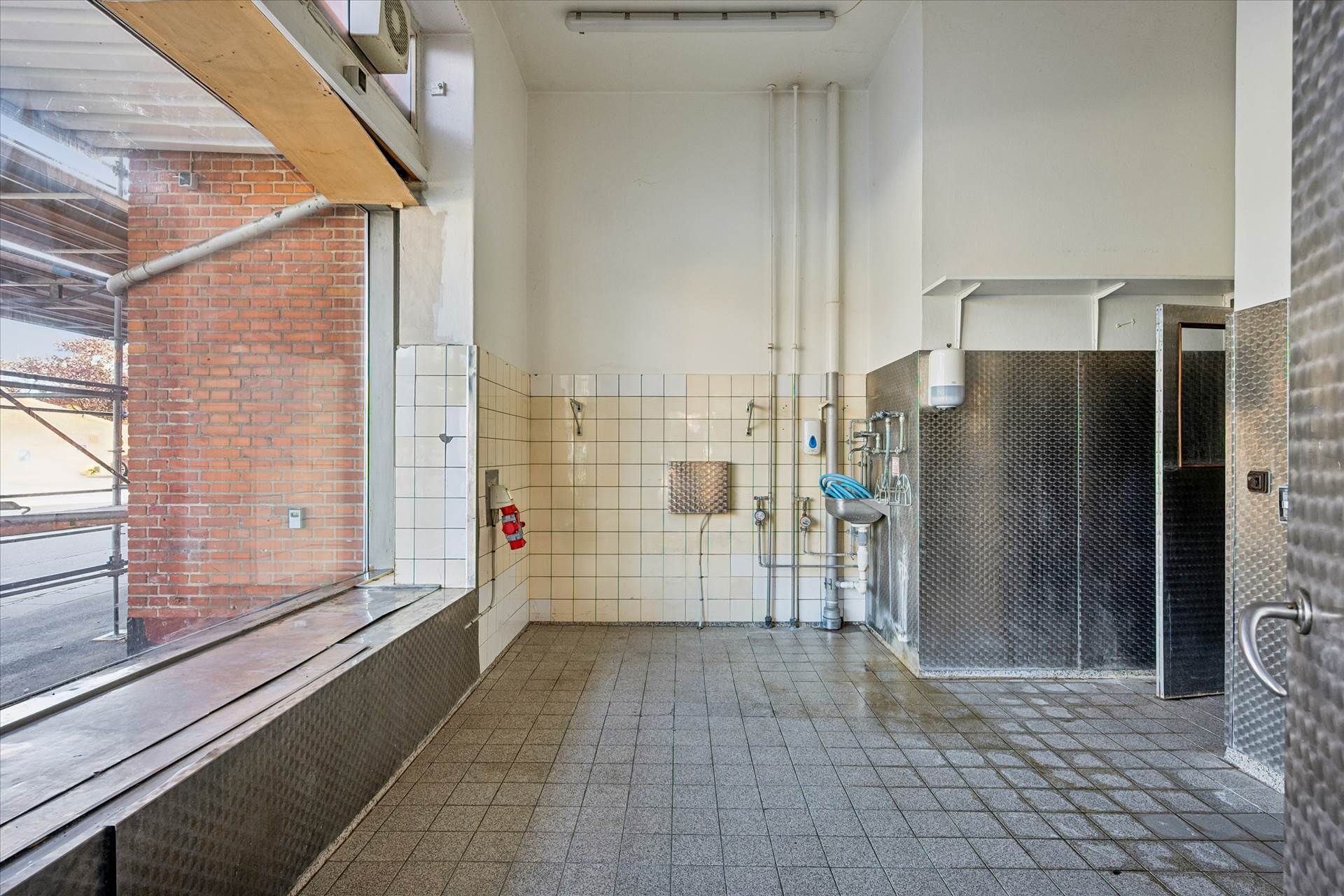 Renseri lokal med rødt murstensvæg, gulvtøj, skraldspande og et rødt brandslukker. På væggen er der fliser, et hængeloft, et