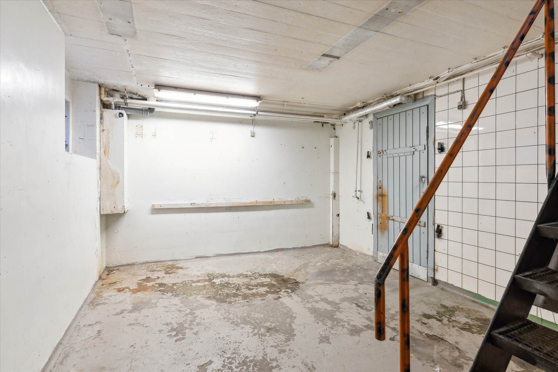 En gammel trappe fører op til en hvid, tom, fugtig kælder med et hul i væggen og en række hvide kakel.