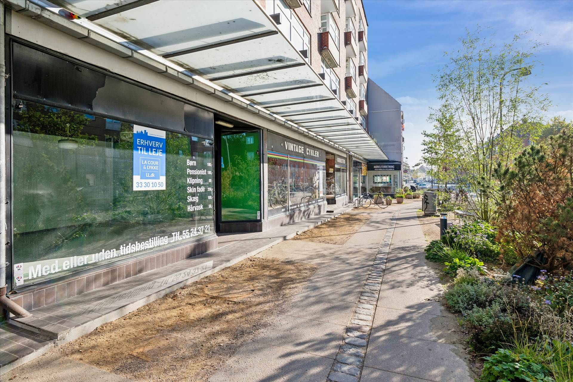 Butik med skiftende reklame, tæt på Vordi og Velo Vintage, udendørs cykelstilling, fortov med grønne planter og parkerede