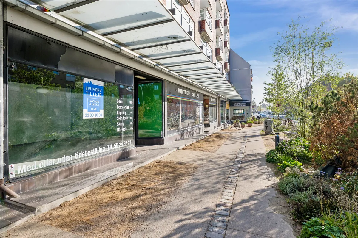 Butik med skiftende reklame, tæt på Vordi og Velo Vintage, udendørs cykelstilling, fortov med grønne planter og parkerede
