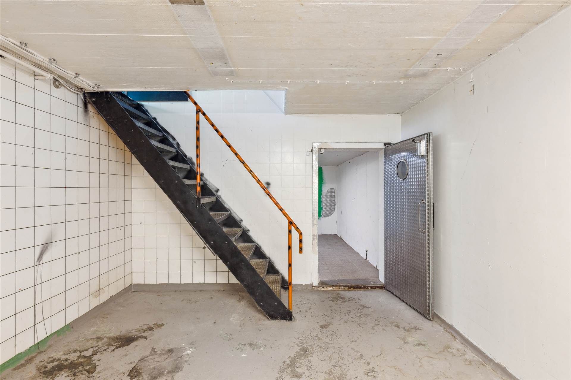 En tom kælder med en trappe, metaldør og en lyse åbning i loftet, begge vægge er hvide og fugtige