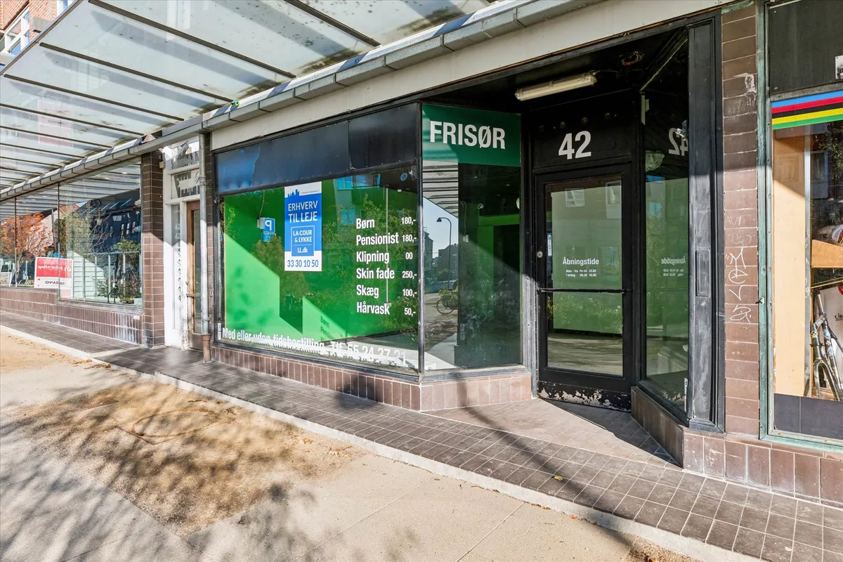 Butiksfront med glasdør og vinduer og skilt med teksten Frisør og åbningstider. På vinduet står der skrift med priser for