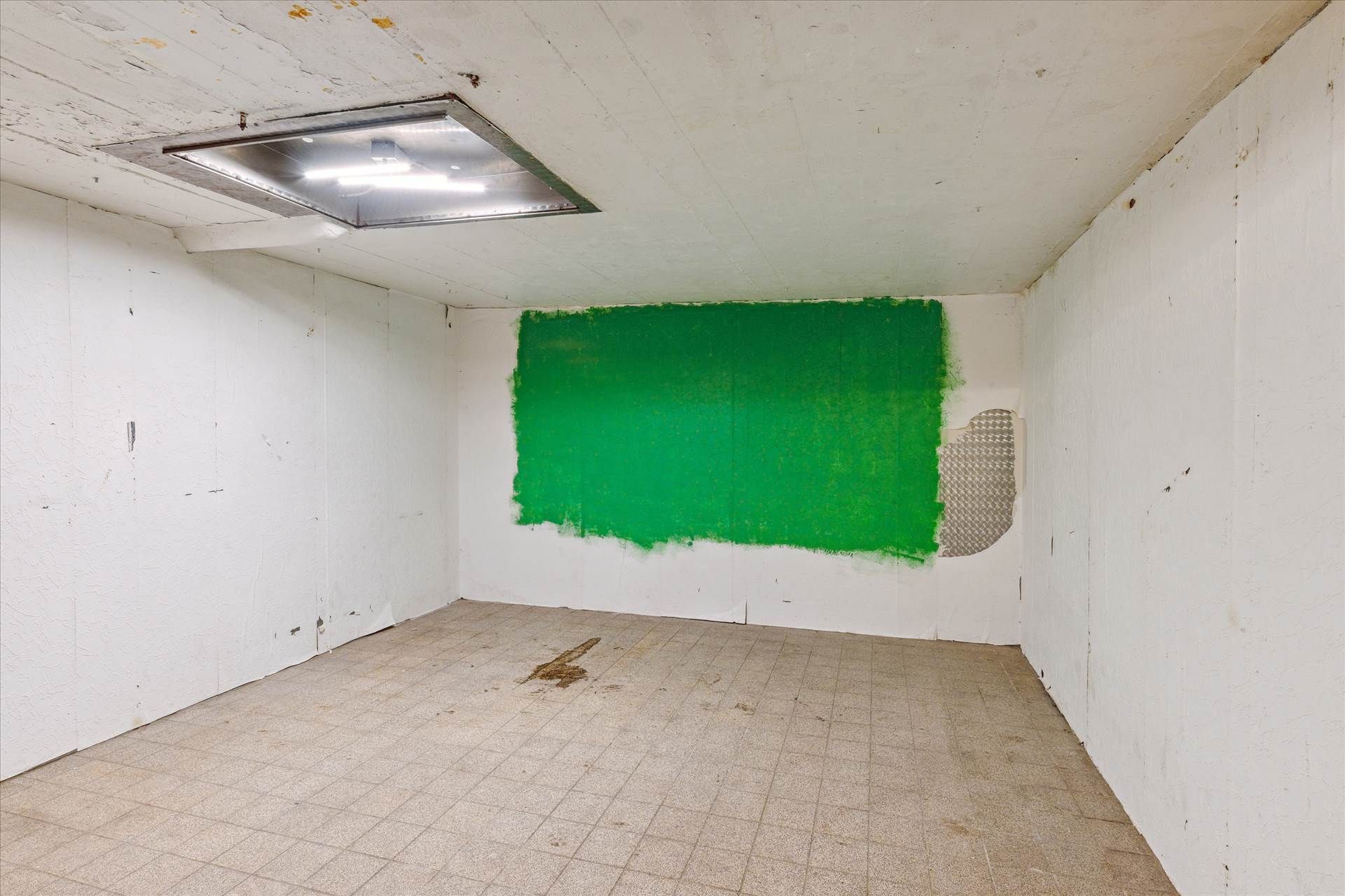 Et tomt rum med hvide vægge og en grøn malingsplet på en af væggene. Gulvet er fliselagt og har snavs på.