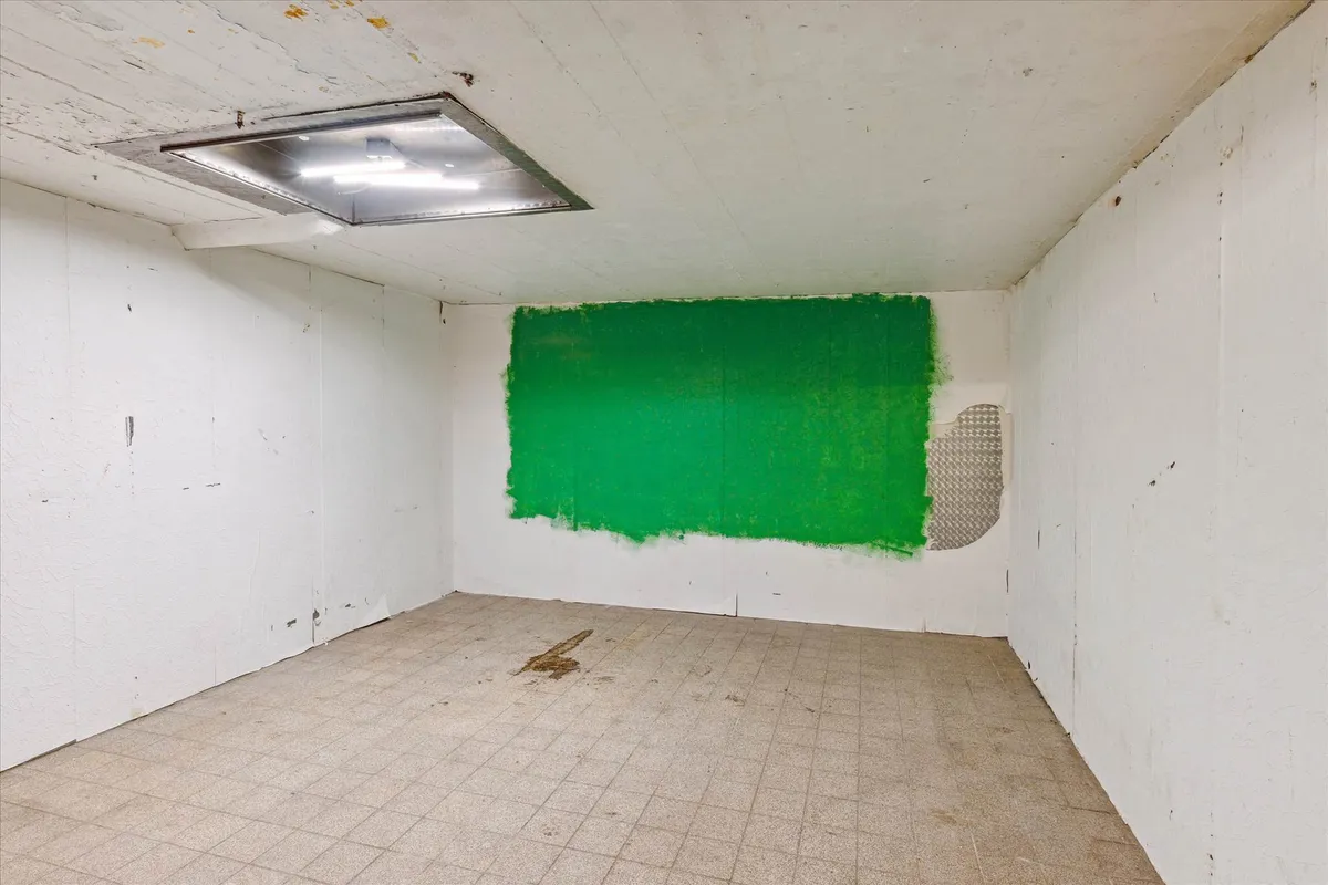 Et tomt rum med hvide vægge og en grøn malingsplet på en af væggene. Gulvet er fliselagt og har snavs på.