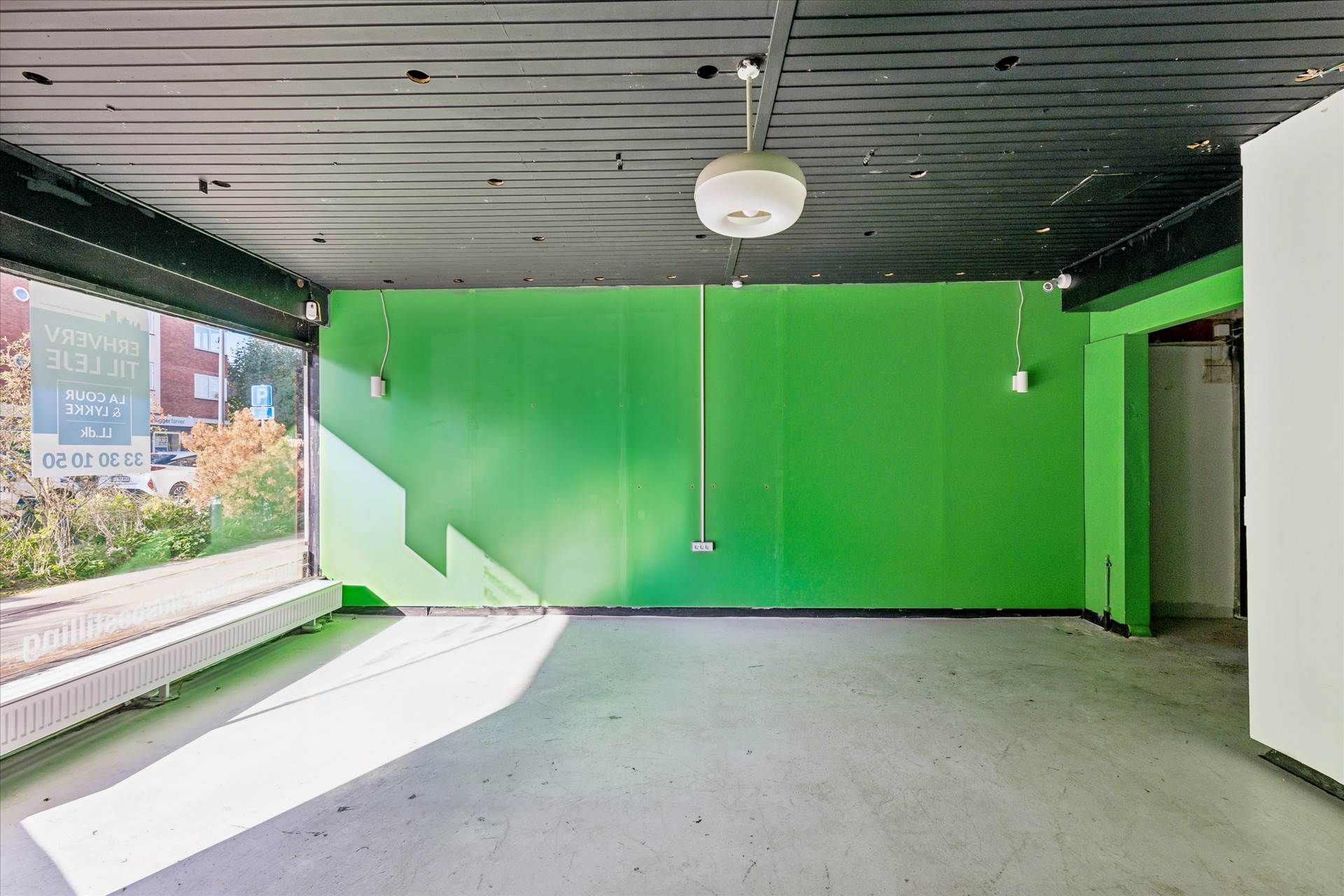 Lokaler til leje med store vinduer og grønne vægge, møbler og udstyr til venstre, solstråler på gulvet, to lysekroner og et