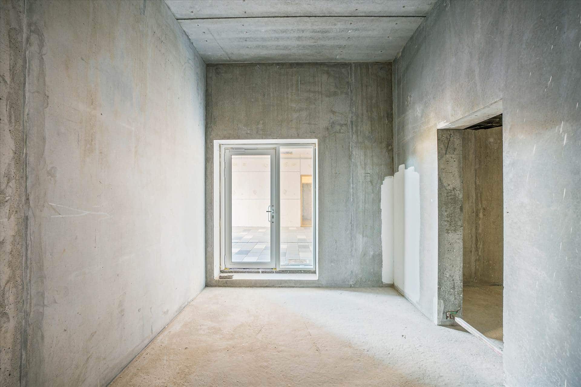 Tomt værelse med hvide vægge og cementgulv samt en stor rude med en dør, der fører til en anden rum.