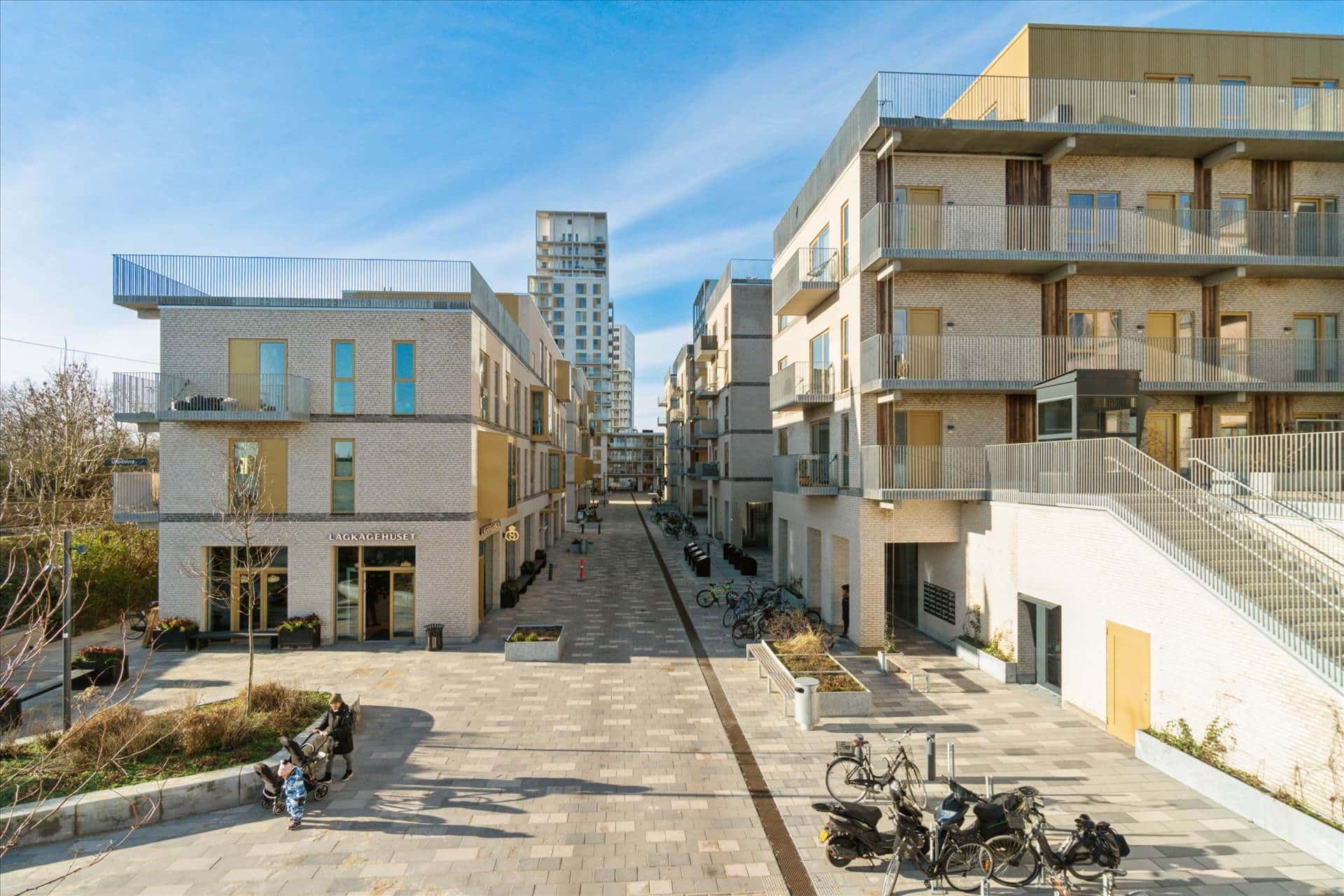 En gågade med butikker, træer og parkeringspladser for cykler og skotere. Et familiepar går med barnevogn, og en person går