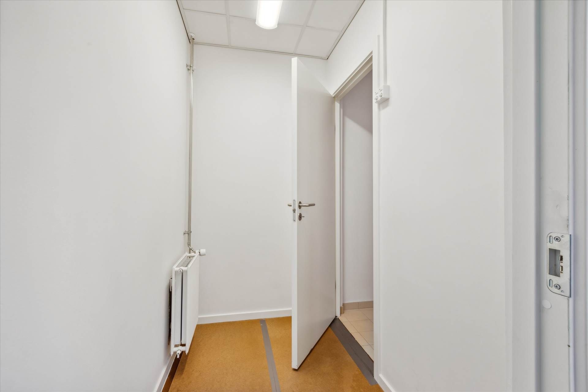 En hvid og tom gang med åben dør, hvor der er et lys over hovedet og en radiator på væggen til venstre