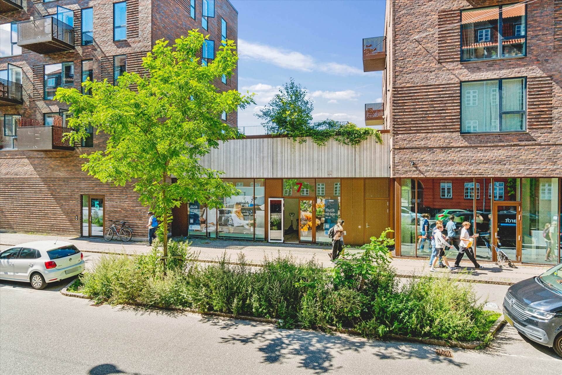 Folk går på gaden med træer og buske. En sølv Volkswagen er parkeret, og der er også en bil med nummerplade. En person er på