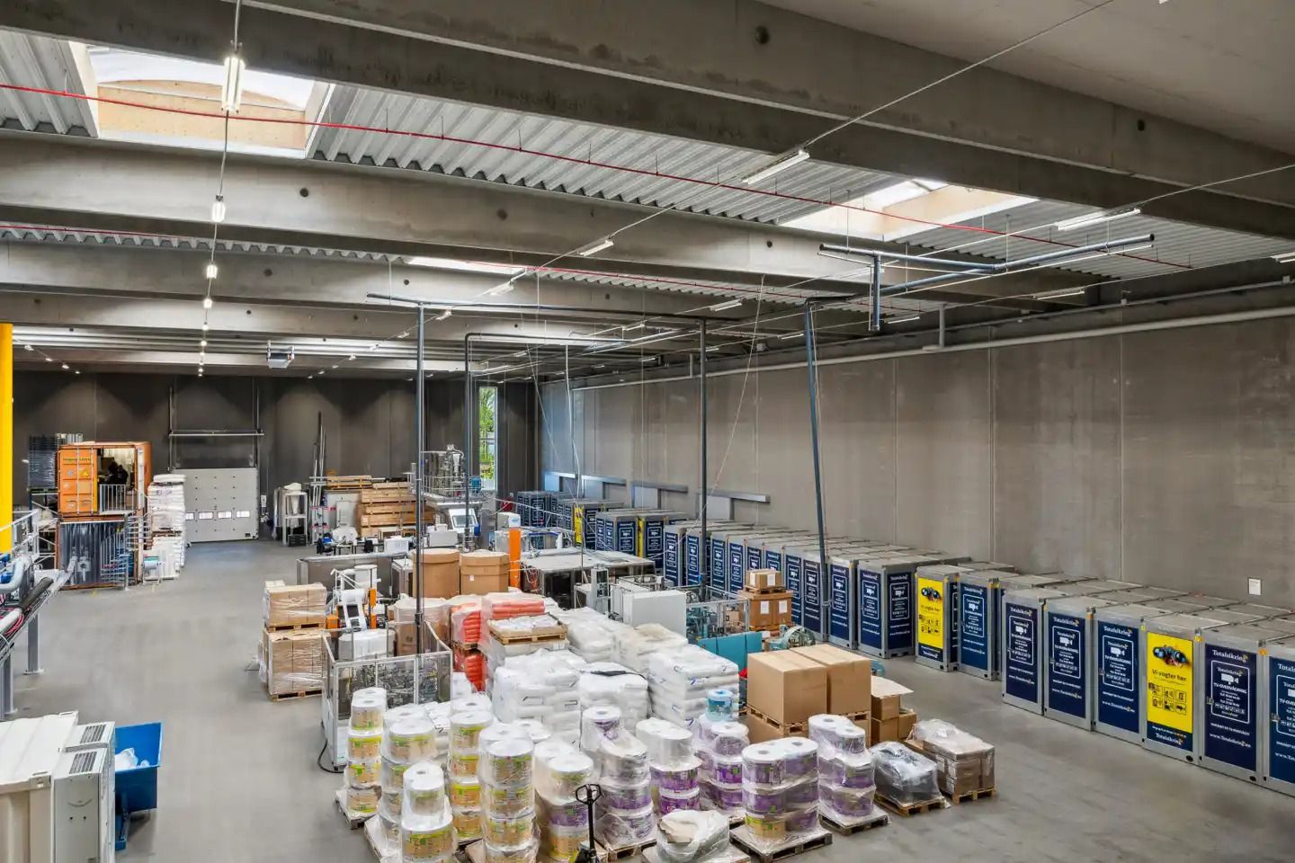 Lager med forskellige slibe- og poleringsmaterialer, papkasser og ruller på paller. Sorte, hvide og gule vægge og gulve