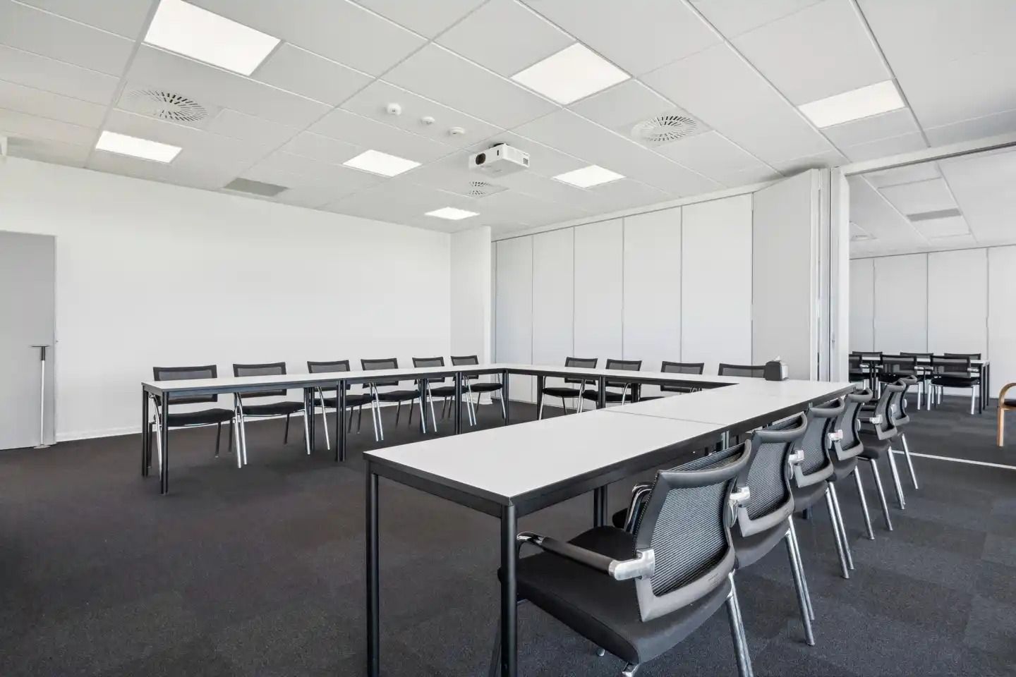 Moderne mødelokale med lange borde og sæder. Hvide vægge og loft med indbygget projektor og ventiler. Gulvet er dækket med