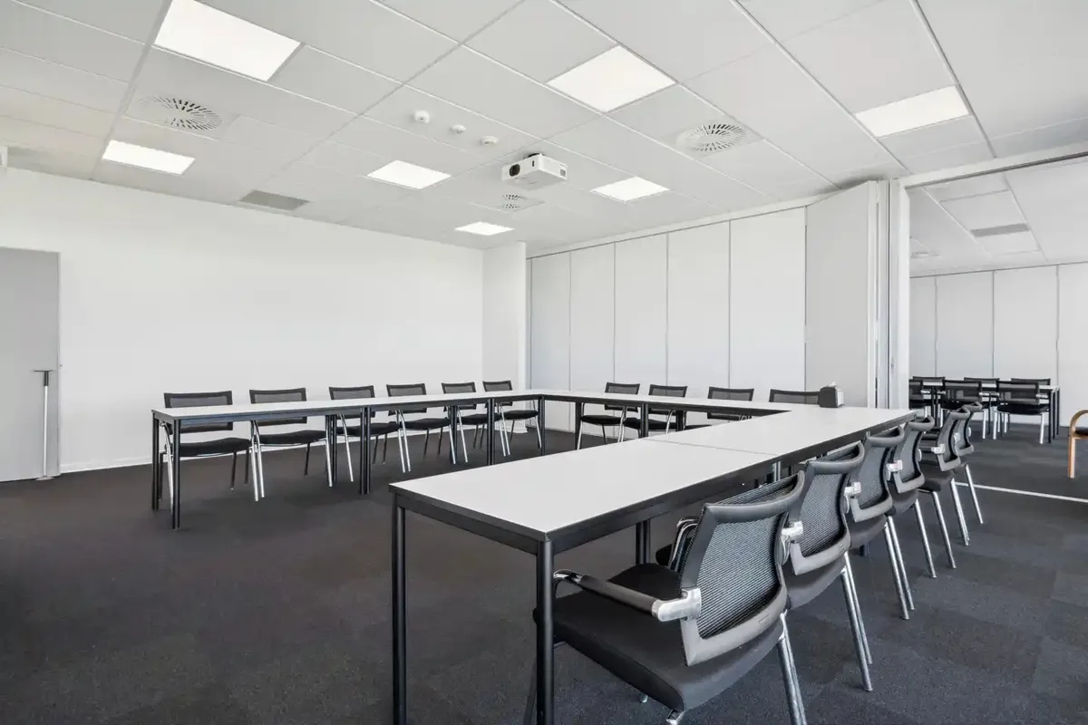 Moderne mødelokale med lange borde og sæder. Hvide vægge og loft med indbygget projektor og ventiler. Gulvet er dækket med