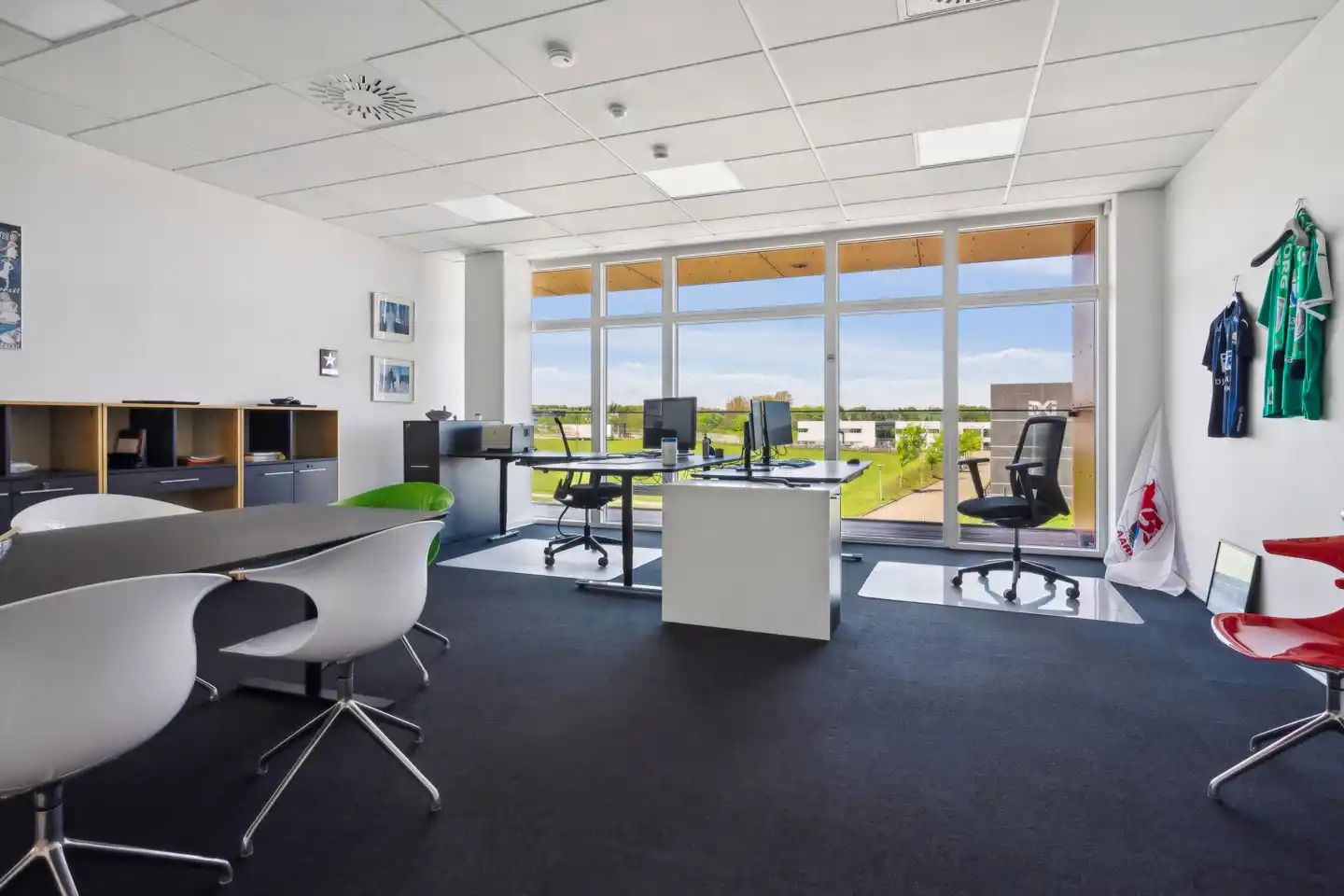 Et moderne kontor med store ruder, højt til loftet og møbler med skarpe linjer samt flere hvide stole og to borde med