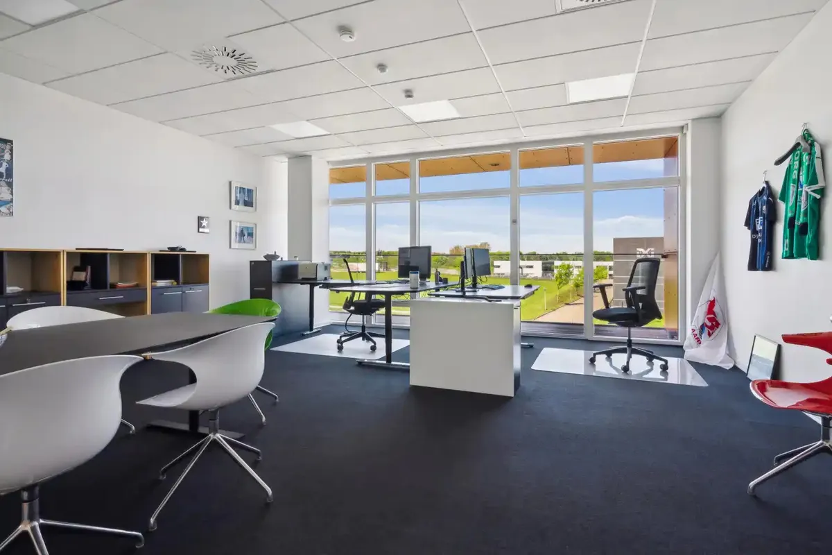 Et moderne kontor med store ruder, højt til loftet og møbler med skarpe linjer samt flere hvide stole og to borde med