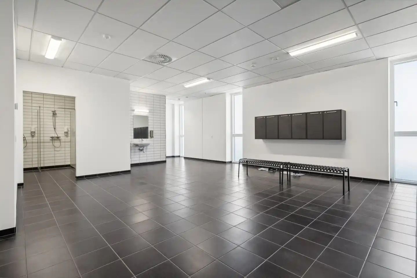 Rum med sort gulv og hvide vægge, med et badeværelse, en spejl, en bænk og flere skabe i hjørnet