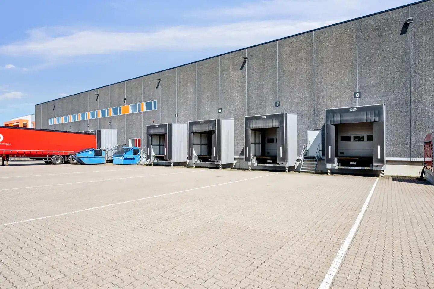 Et stort, gråt lagerhus med åbne portdøre og en stor rødslere parkeret foran
