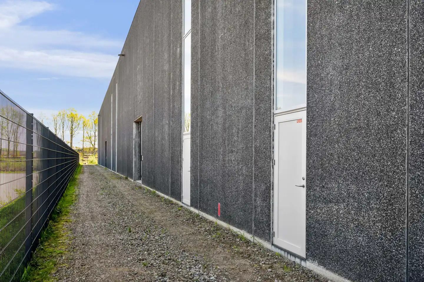 En stor lagerbygning med stålporte og cementskraldebelægning. Bygningen er omgivet af en høj metalhegn. Der er to hvide