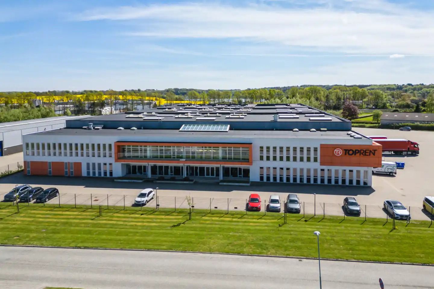 En fabrik med solceller på taget og en fortovsparkering foran. Der er køretøjer og busser parkeret, og det er et lyst og