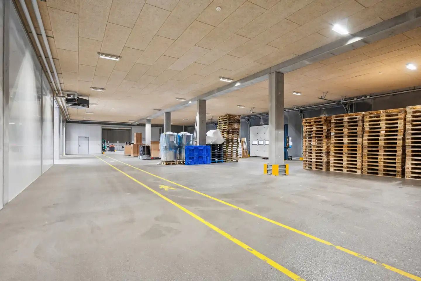 Lager med højloft, flere rækker af træpæle og to høje gulvmonterede søjler. Gulvet er markeret med gule striber.