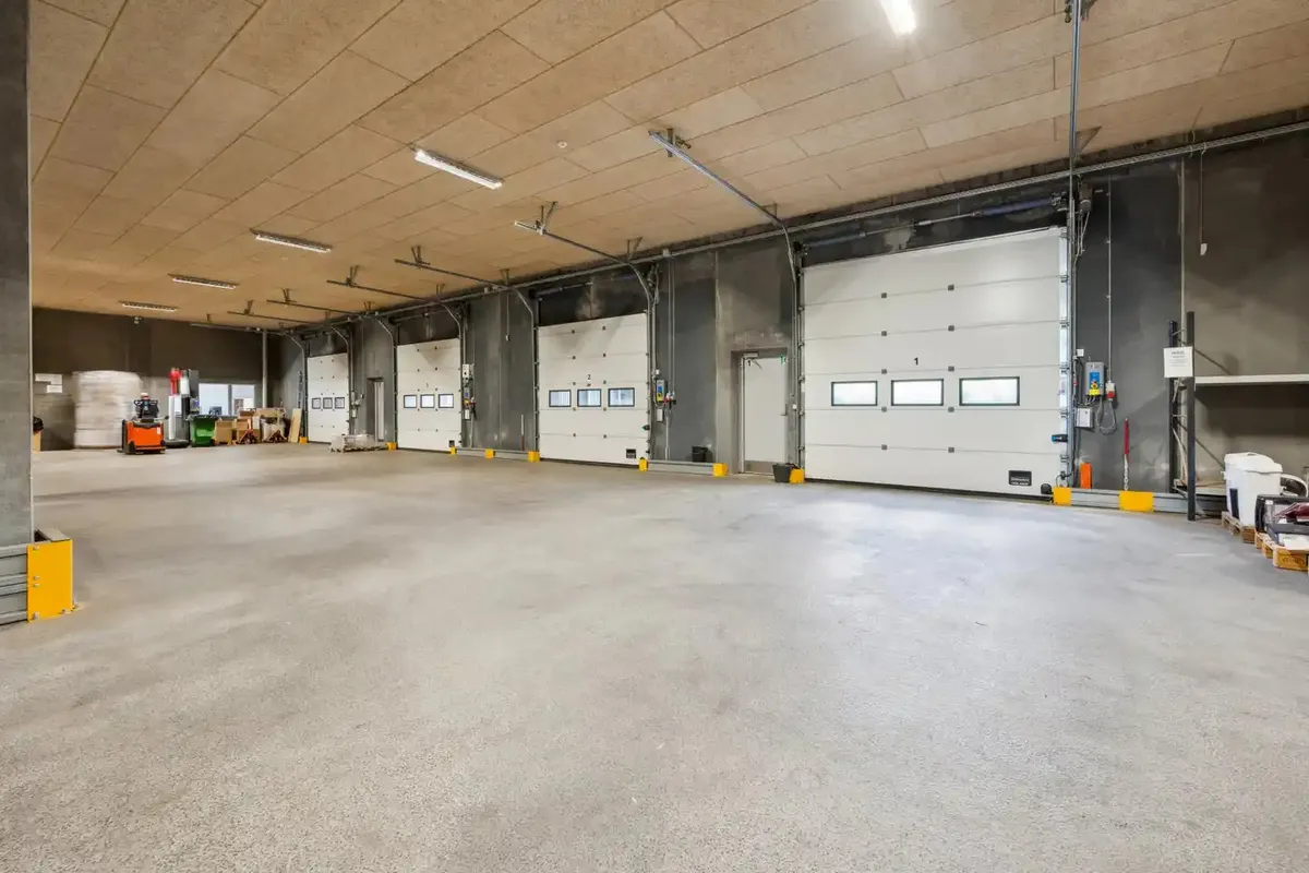 En stor, tom bygning med højt til lofts og hvide garageporte samt gul gulvmærkning og røde og hvide søjler