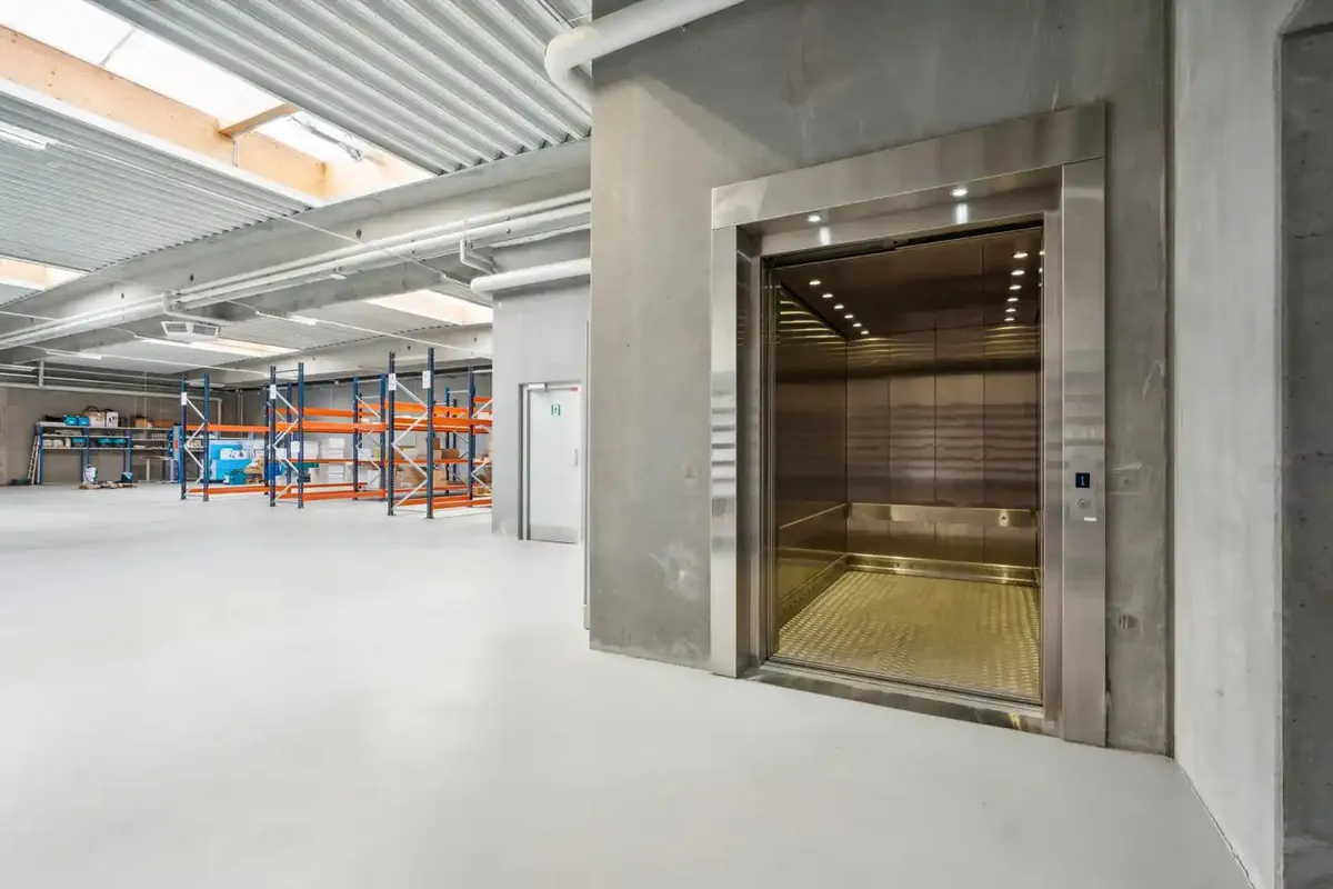 Lager med store mængder varer og flere reoler. Metal elevator og port i den ene side. Lyse lofter og hvide vægge.