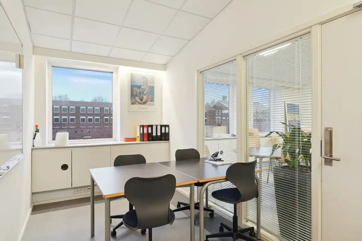 Kontorlokaler med bord, stole, tavle, kunstværk, plante og vinduer med gardiner og udsigt til byen