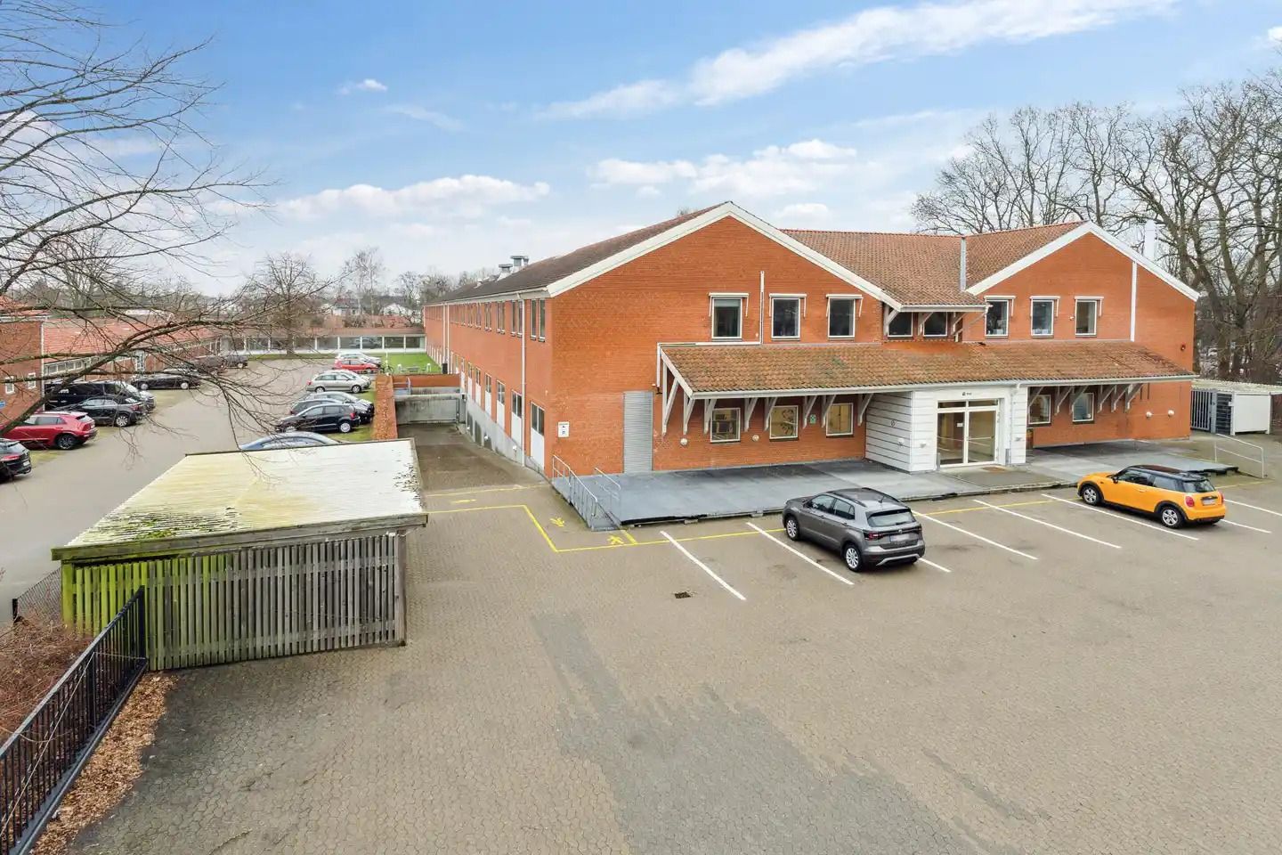 Rødt byggeværk med mange vinduer og garagedør samt parkeringsplads med flere parkerede biler og en stigning til en entrence