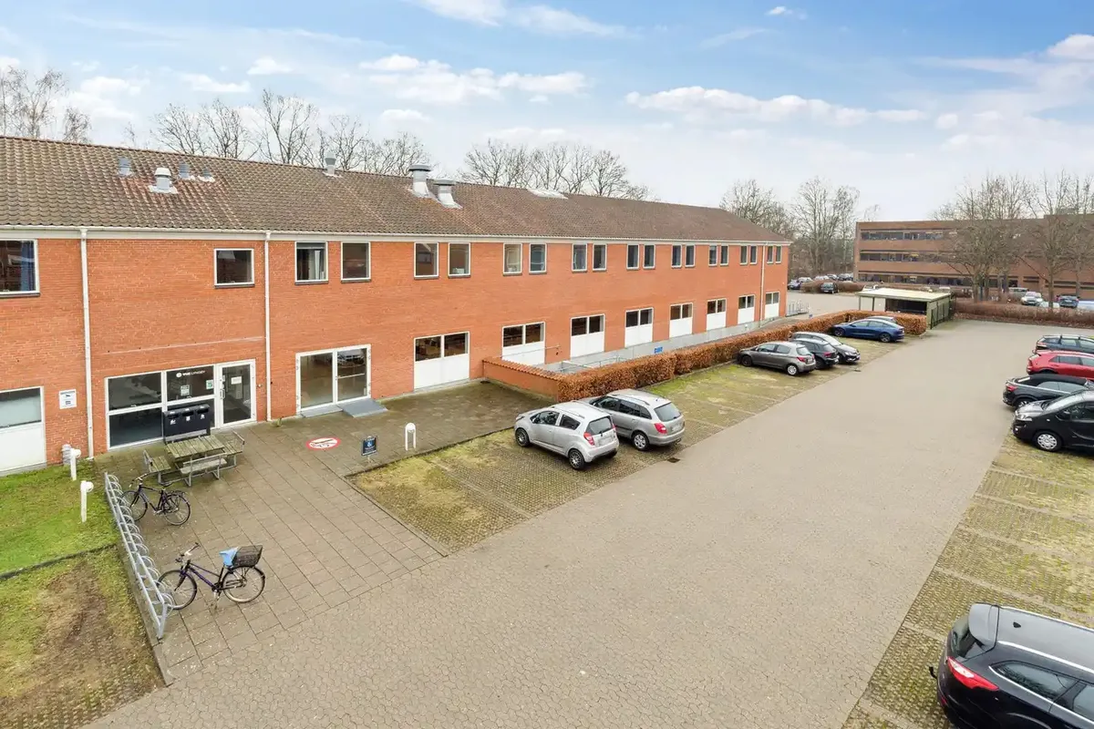 To parkeringspladser med flere parkerede biler og enkelte cykler foran et bygningskompleks med flere lejligheder og