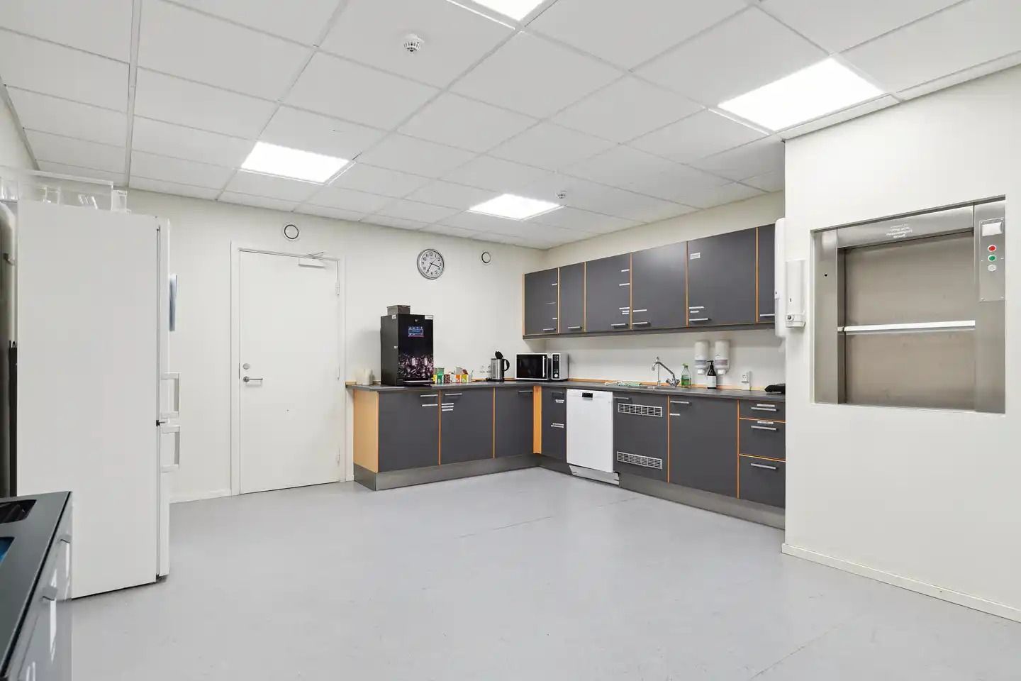 Et køkken med hvidt loft og gulv samt grå og brune skabe og indbyggede apparater som køleskab, mikroovn og vaskemaskine