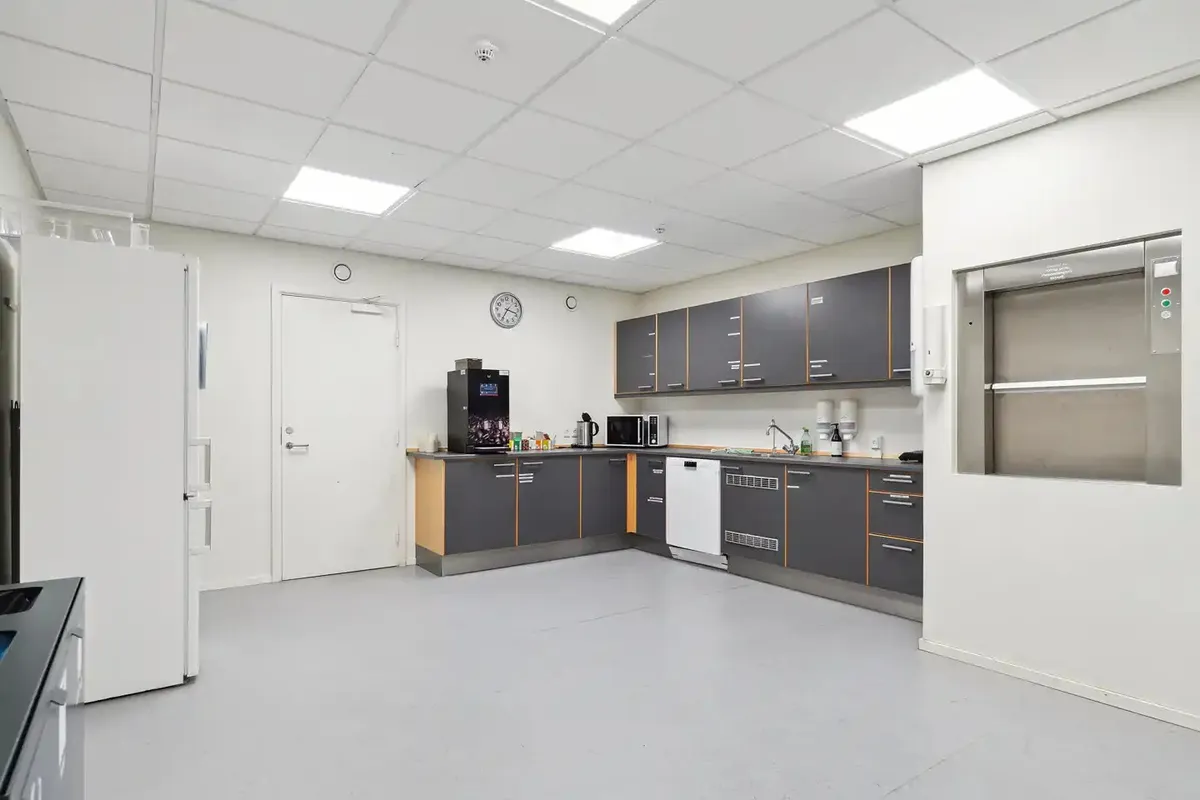 Et køkken med hvidt loft og gulv samt grå og brune skabe og indbyggede apparater som køleskab, mikroovn og vaskemaskine