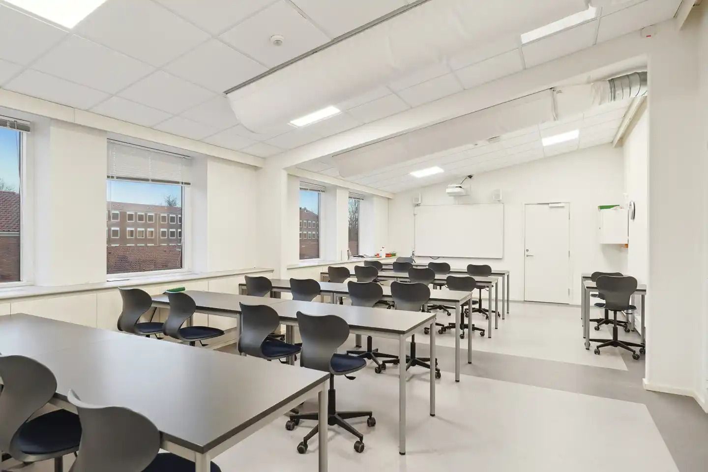 En tom klasseværelse med lysegul væg og gulvtæppe, flere skriveborde og stole, højt hængende projektor og hvide loftslampe