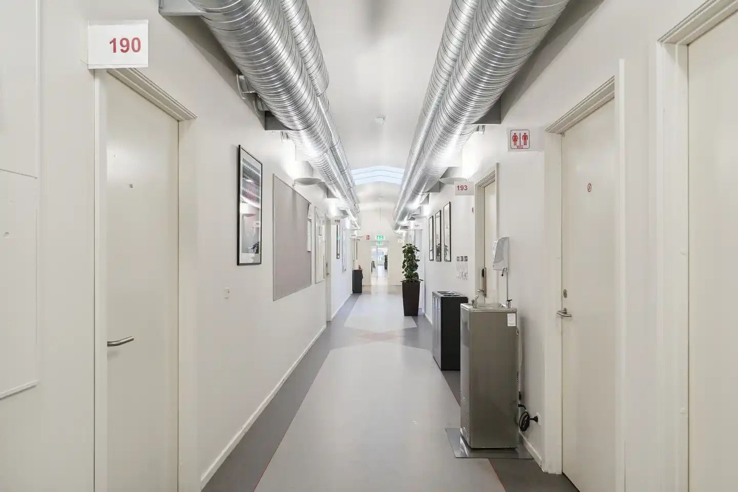 En række hvide døre og rummelige gange i en bygning med lyse lofter og gulvbelægning