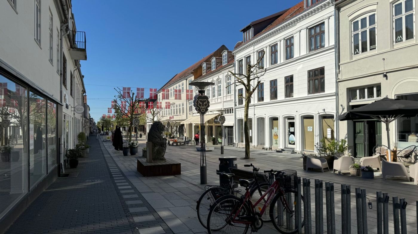 Gaden med hvide bygninger og terrasser, udsmykket med flag og statuer, og to cykler parkeret ved siden af en metalbøss.