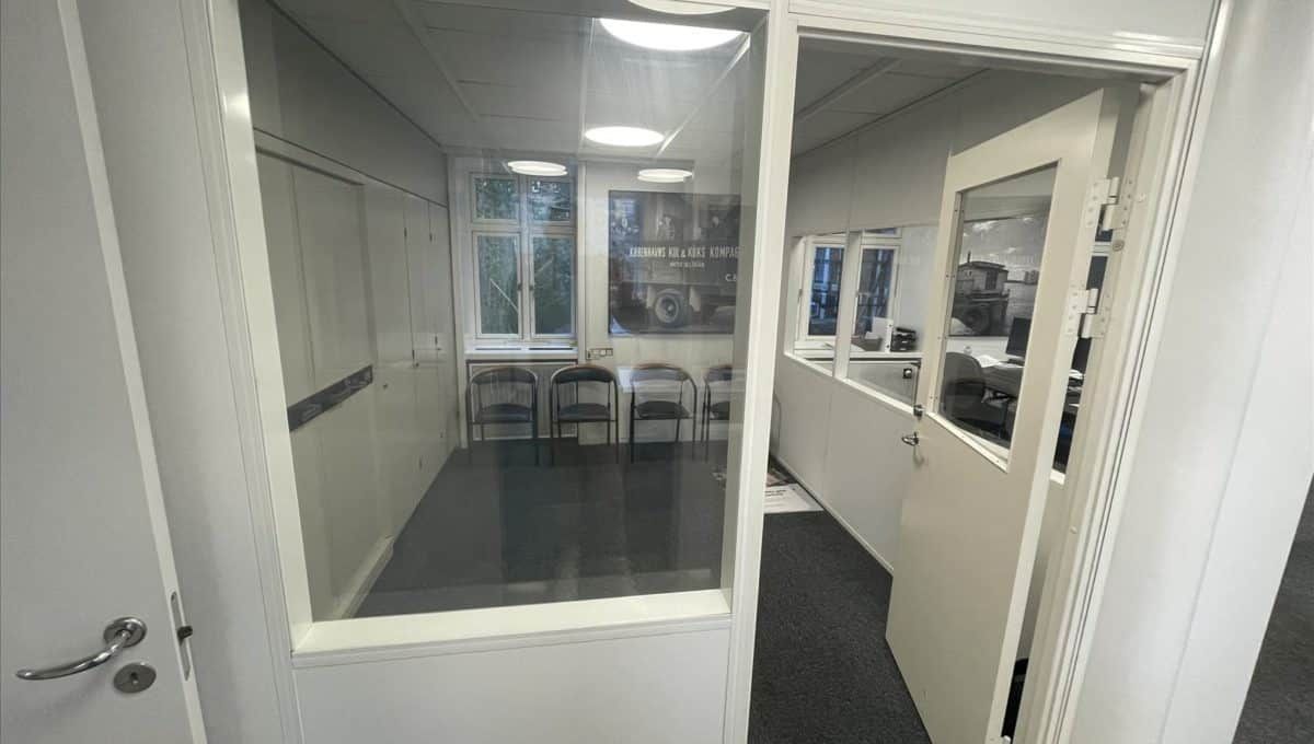 Glassdør med håndtag og åbne indgangsdøre til et moderne kontor med hvide vægge og lyse loftslamper