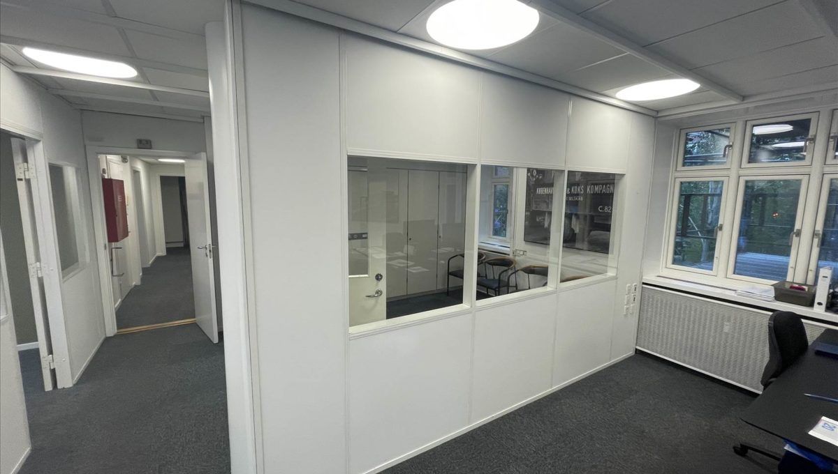 Kontor med hvide vægge, højt til loftet og store vinduer med udsigt til gårdhaven og grønne træer.