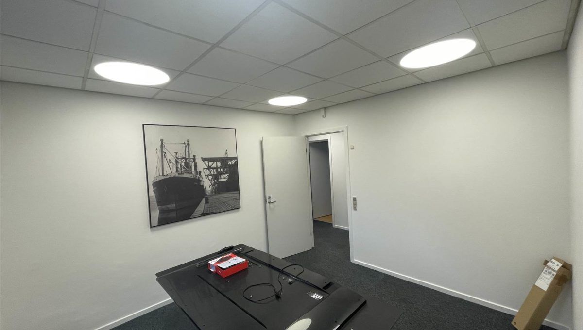 Kontorlokaler med lyse hvide vægge og loft. En sort skrivebord med to røde bokse og en kabel. Et billede hænger på væggen og