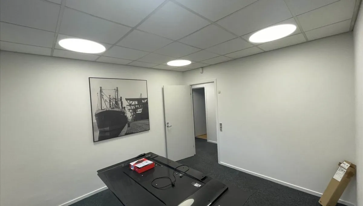 Kontorlokaler med lyse hvide vægge og loft. En sort skrivebord med to røde bokse og en kabel. Et billede hænger på væggen og